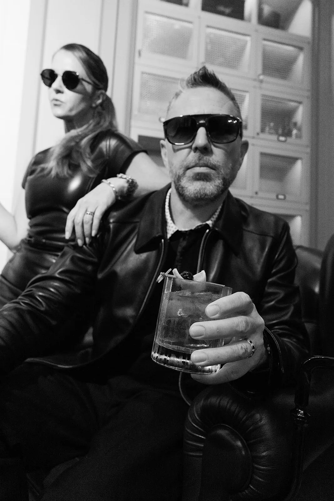 Black and white portrait of a man holding a cocktail, with a woman behind him in a leather outfit