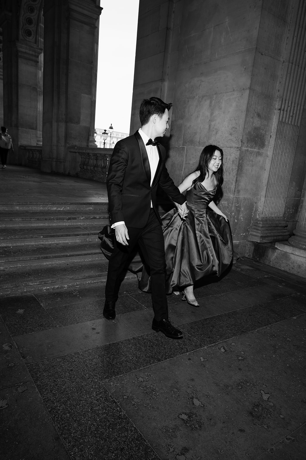 Black and white editorial couple walking beneath the Louvre arcades in Paris