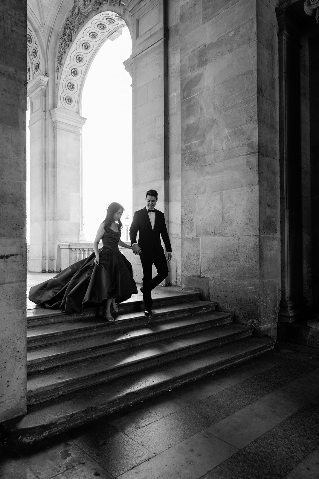 Black and white pre-wedding couple descending the monumental Louvre staircase in Paris