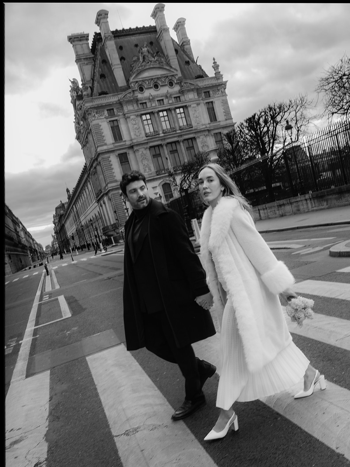 Black-and-white editorial couple crossing the street beside the Louvre Museum in Paris