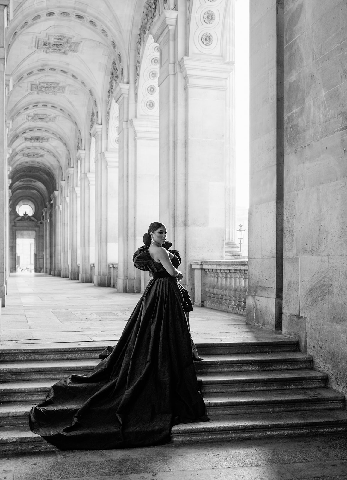 Black-and-white portrait of Candice Craig on the Louvre steps wearing a sculptural black gown
