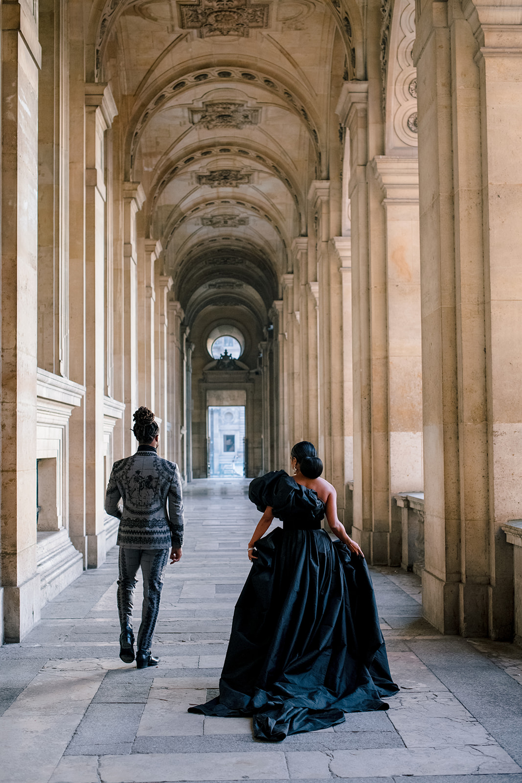 Candice Craig and Omar Bolden walking together beneath the Louvre arcades in Paris