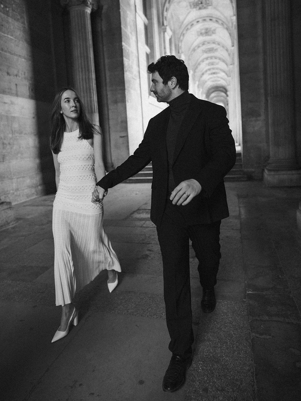 Black-and-white editorial couple walking beneath the Louvre colonnade in Paris