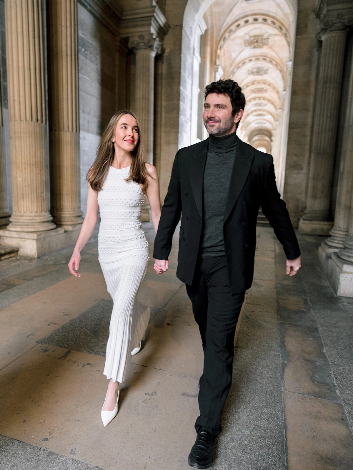 Fashion-forward couple walking beneath the Louvre colonnade during a Paris photoshoot